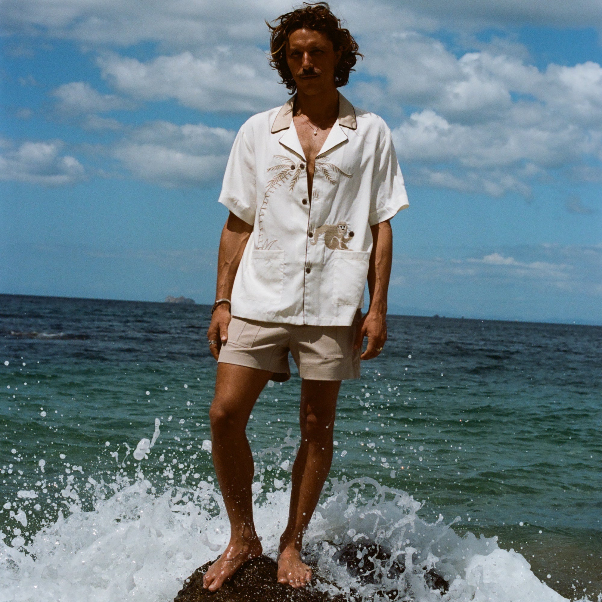 Male model pictured wearing the 'Monkey Business' Shirt with our Khaki Stowaway Shorts. He is standing on a rock at the beach with a wave splashing behind him.