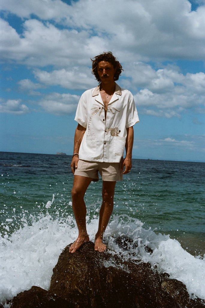 Male model pictured wearing the 'Monkey Business' Shirt with our Khaki Stowaway Shorts. He is standing on a rock at the beach with a wave splashing behind him.