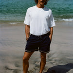 A brunette man in a white terry tee and navy swim shorts standing barefoot on a beach, with his hands in pockets.