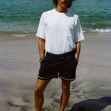 A brunette man in a white terry tee and navy swim shorts standing barefoot on a beach, with his hands in pockets.