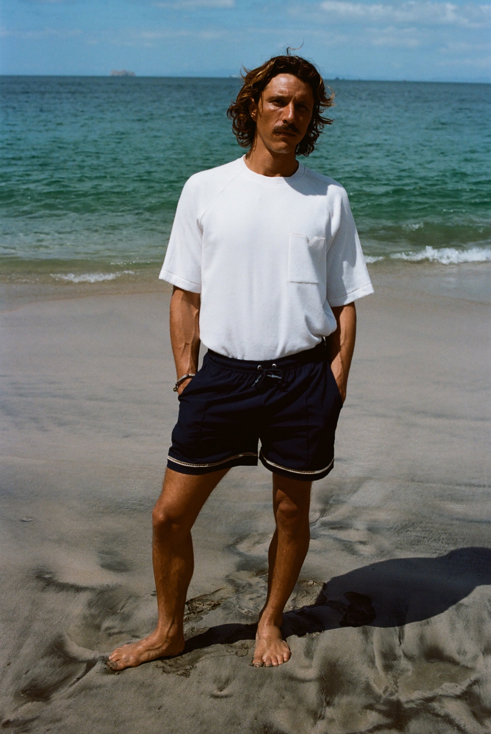 A brunette man in a white terry tee and navy swim shorts standing barefoot on a beach, with his hands in pockets.