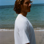 Side image of a brunette man wearing the white terry tee while walking on the beach. 