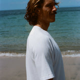 Side image of a brunette man wearing the white terry tee while walking on the beach. 