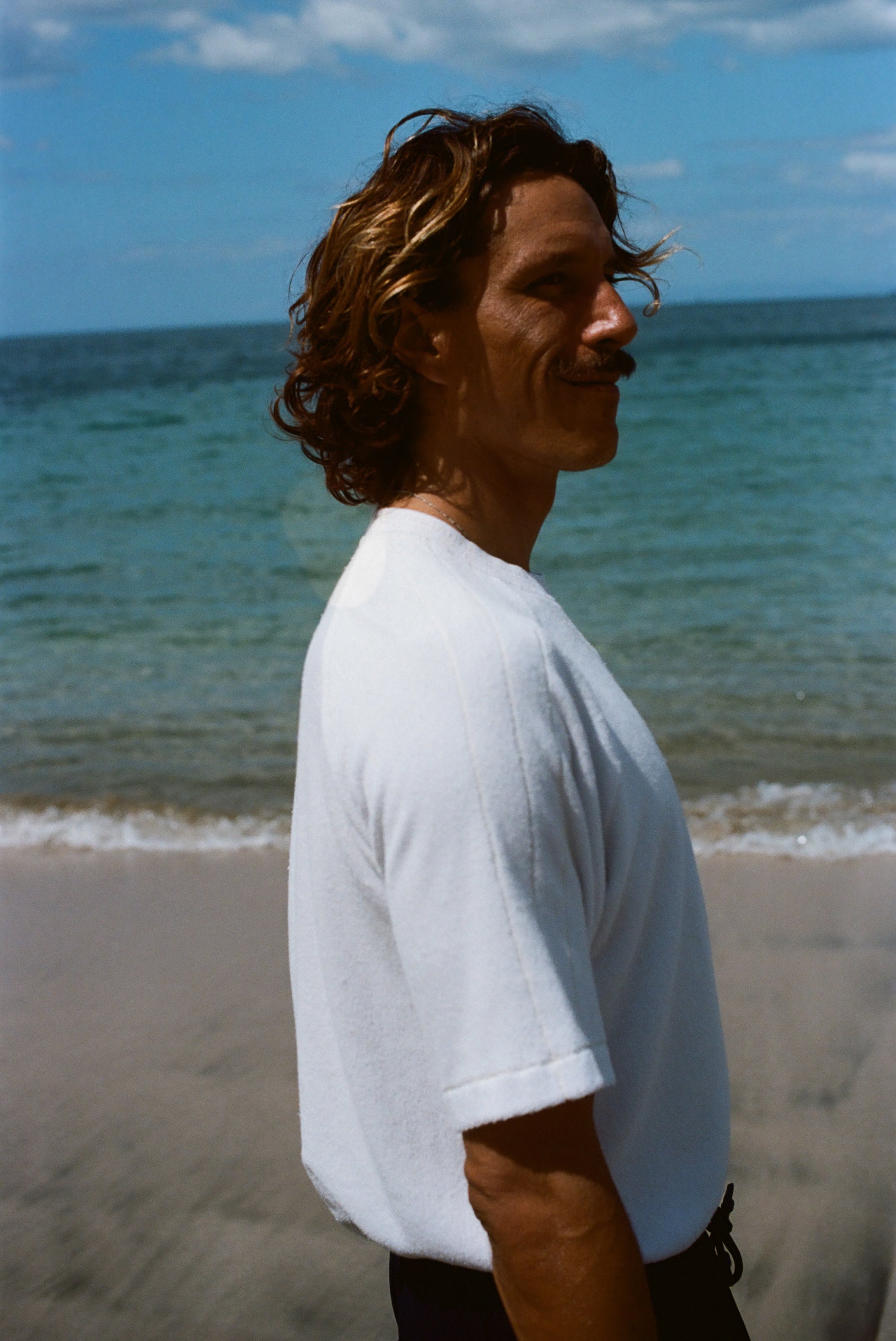 Side image of a brunette man wearing the white terry tee while walking on the beach. 