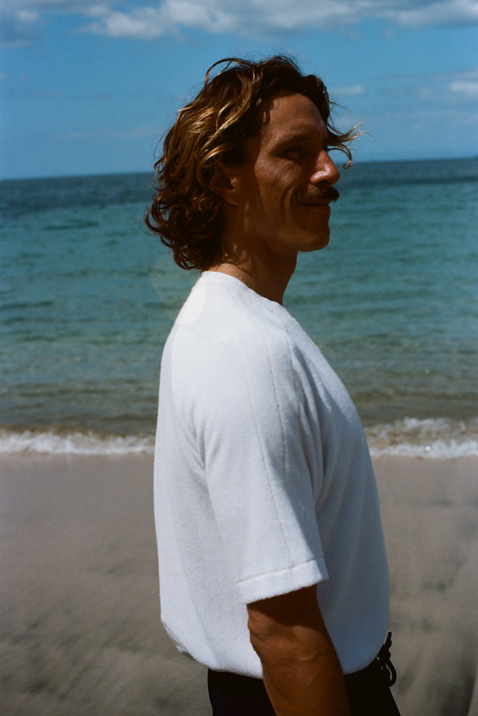 Side image of a brunette man wearing the white terry tee while walking on the beach. 