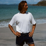 Man standing on beach wearing the white terry t-shirt tucked into a pair of navy swim trunks. His hands are on his hips and he is looking off to the side.