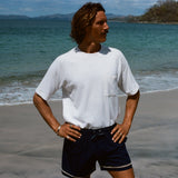 Man standing on beach wearing the white terry t-shirt tucked into a pair of navy swim trunks. His hands are on his hips and he is looking off to the side.