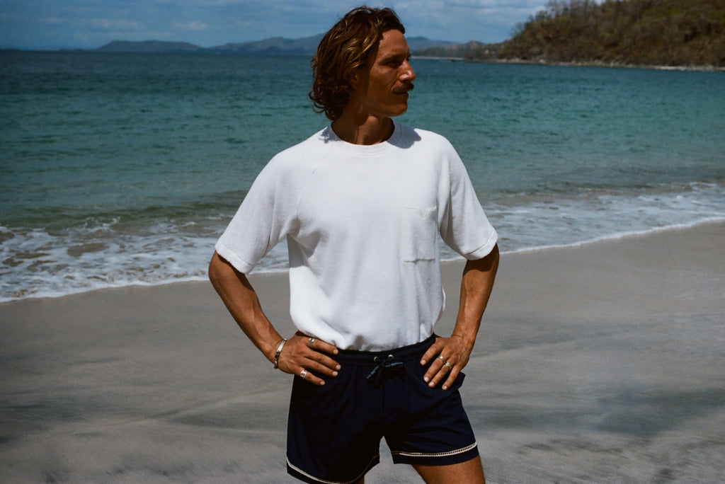 Man standing on beach wearing the white terry t-shirt tucked into a pair of navy swim trunks. His hands are on his hips and he is looking off to the side.