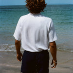 Image from behind of a brunette man wearing our white terry cloth tee with navy swim trunks and looking off into the ocean.