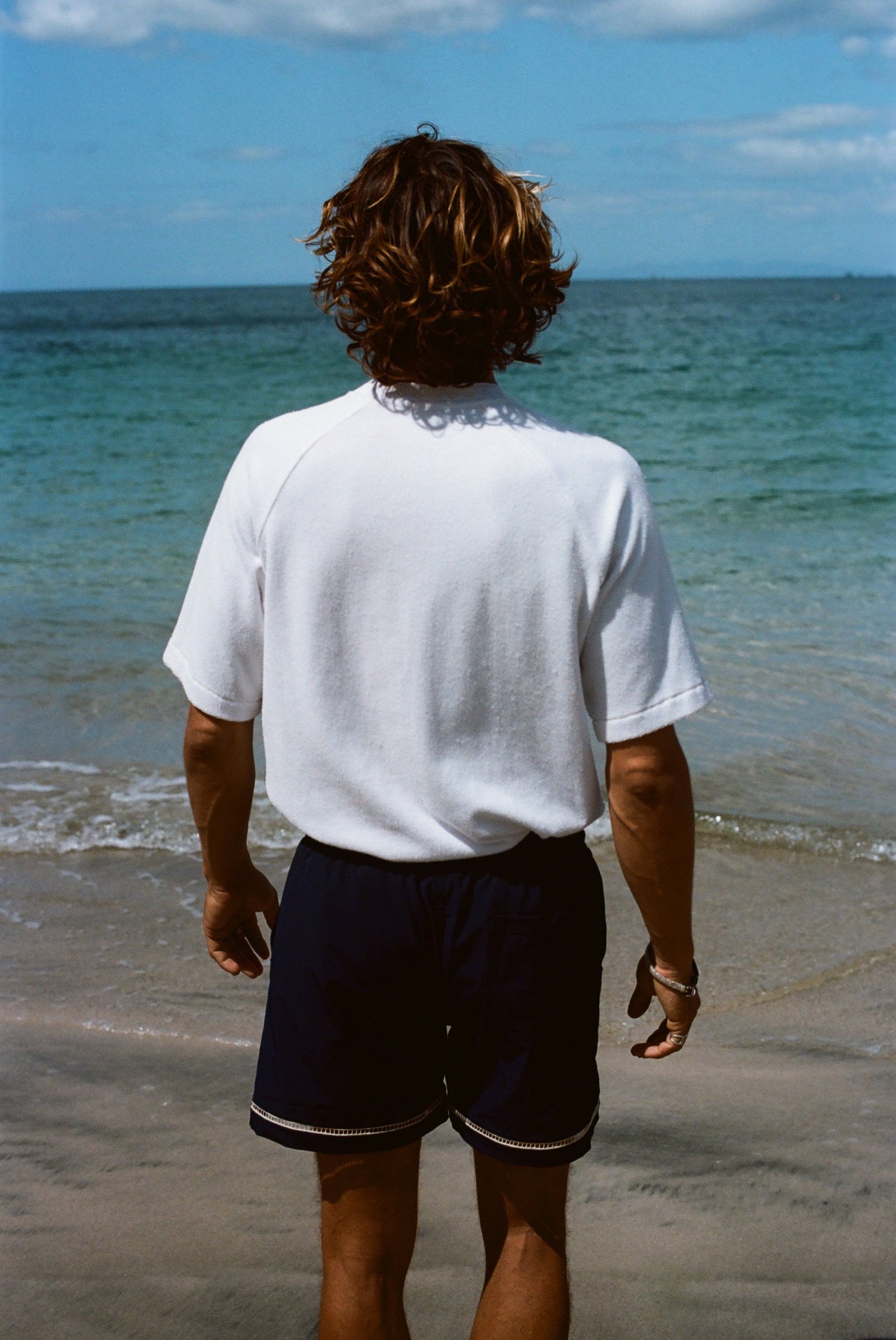 Image from behind of a brunette man wearing our white terry cloth tee with navy swim trunks and looking off into the ocean.