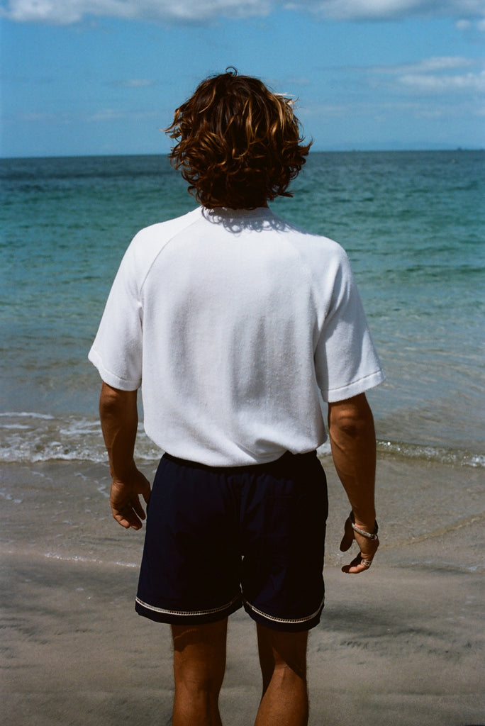 Image from behind of a brunette man wearing our white terry cloth tee with navy swim trunks and looking off into the ocean.