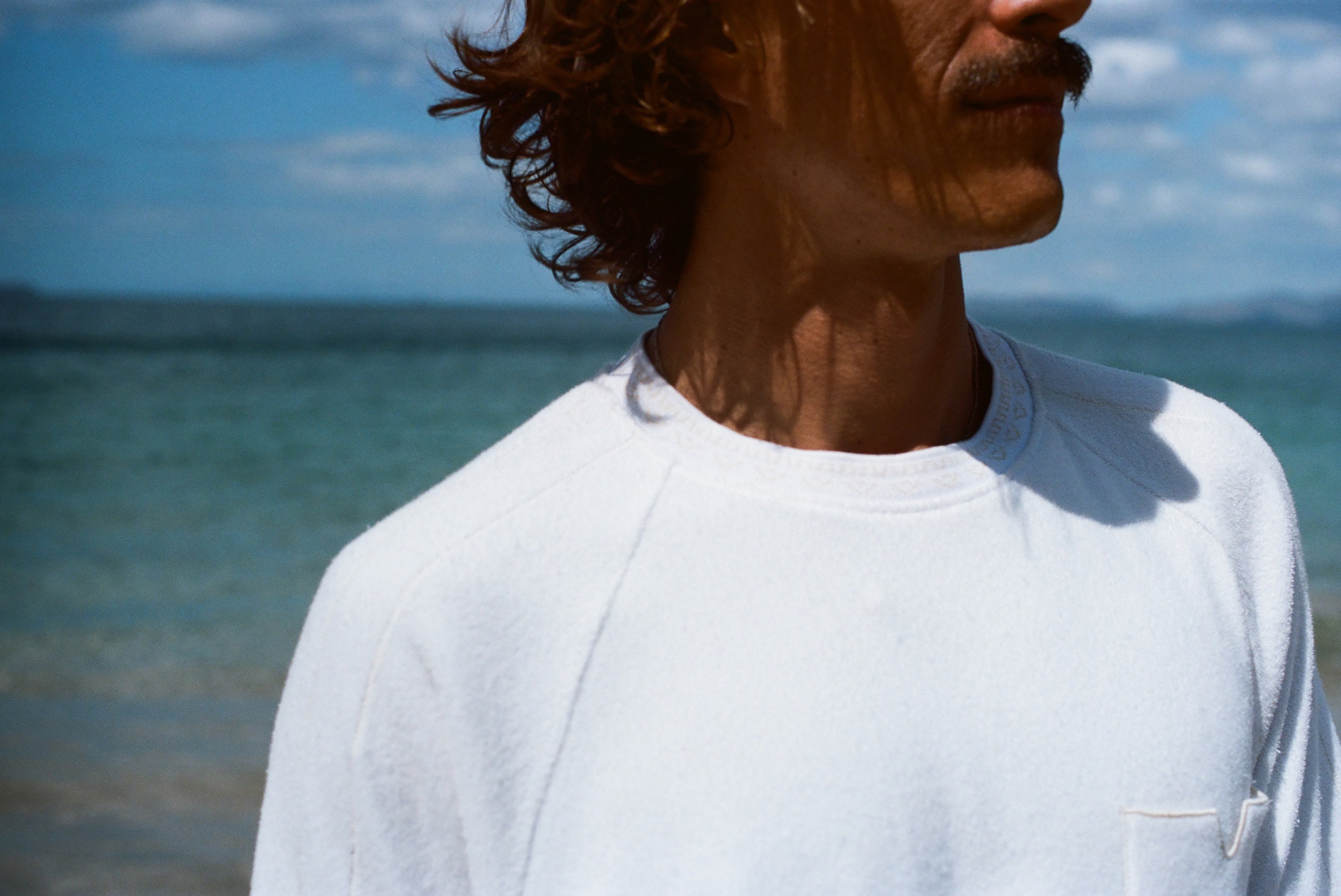 Zoomed in image of the collar of our white terry tee, modeled by a brunette man standing on the beach.