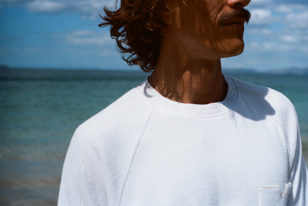 Zoomed in image of the collar of our white terry tee, modeled by a brunette man standing on the beach.