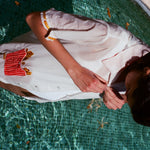 Woman standing in a pool wearing a white short-sleeve button-down shirt with embroidered yellow fries on the sleeves and a red gingham pocket filled with embroidered fries. The person is buttoning the shirt.