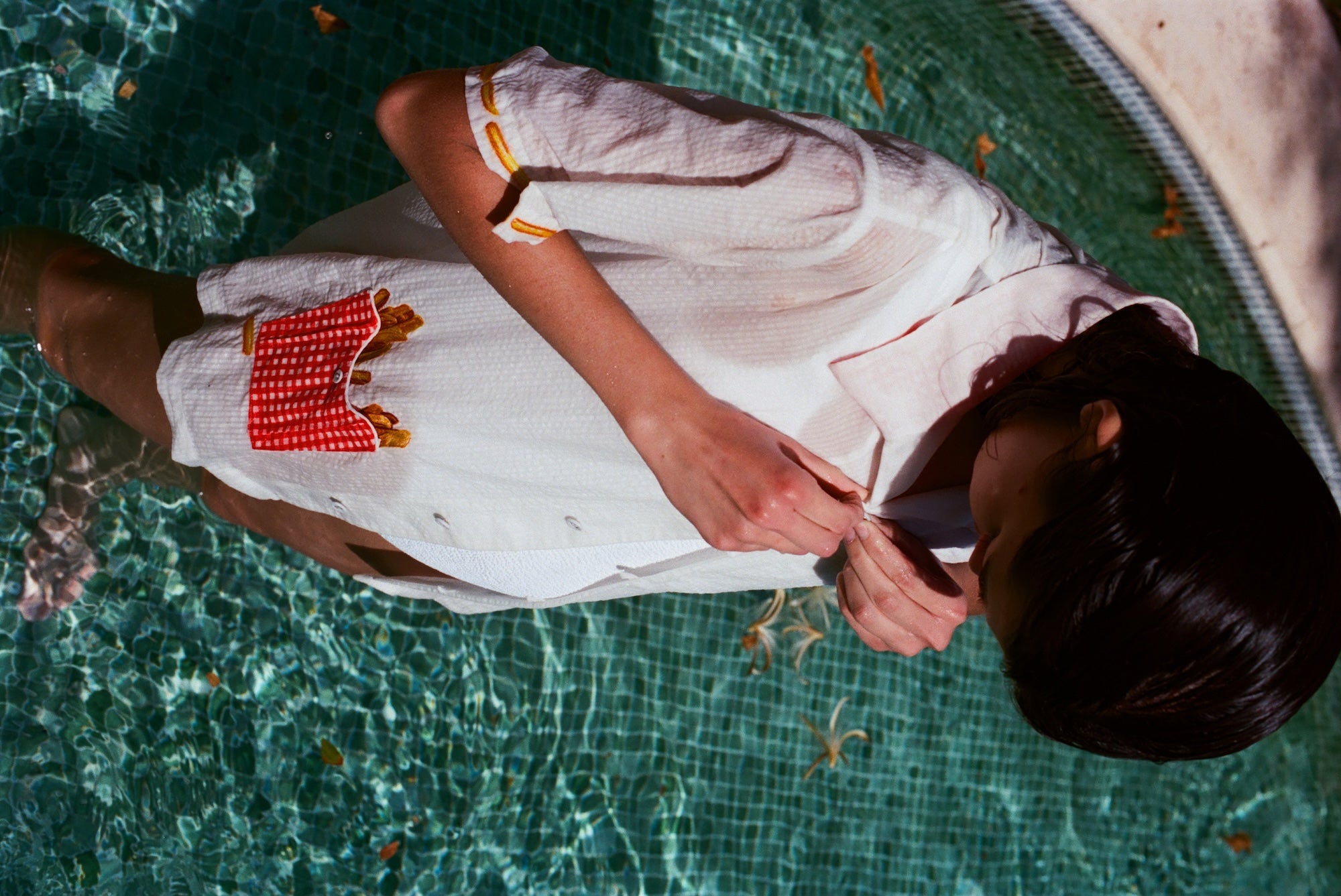 Woman standing in a pool wearing a white short-sleeve button-down shirt with embroidered yellow fries on the sleeves and a red gingham pocket filled with embroidered fries. The person is buttoning the shirt.