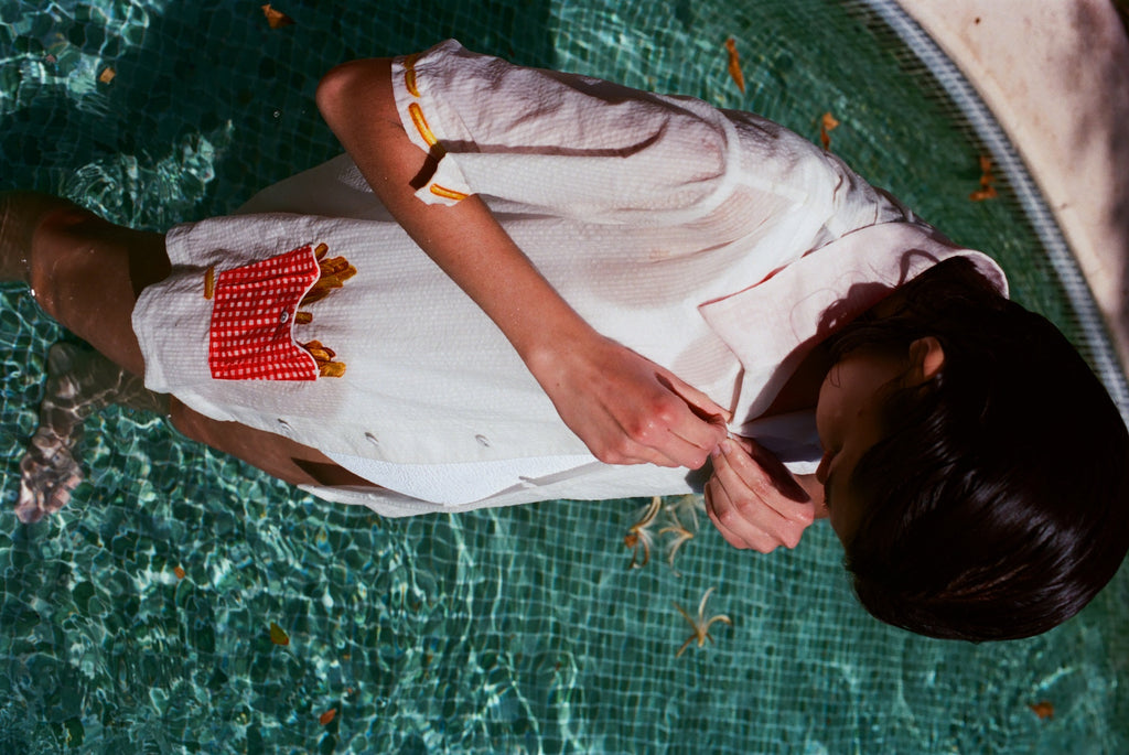 Woman standing in a pool wearing a white short-sleeve button-down shirt with embroidered yellow fries on the sleeves and a red gingham pocket filled with embroidered fries. The person is buttoning the shirt.