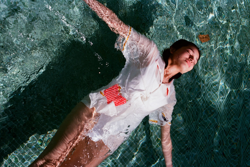 Woman floating in a pool wearing a white button-down shirt with embroidered fries in a red gingham pocket and fry detailing on the sleeves.
