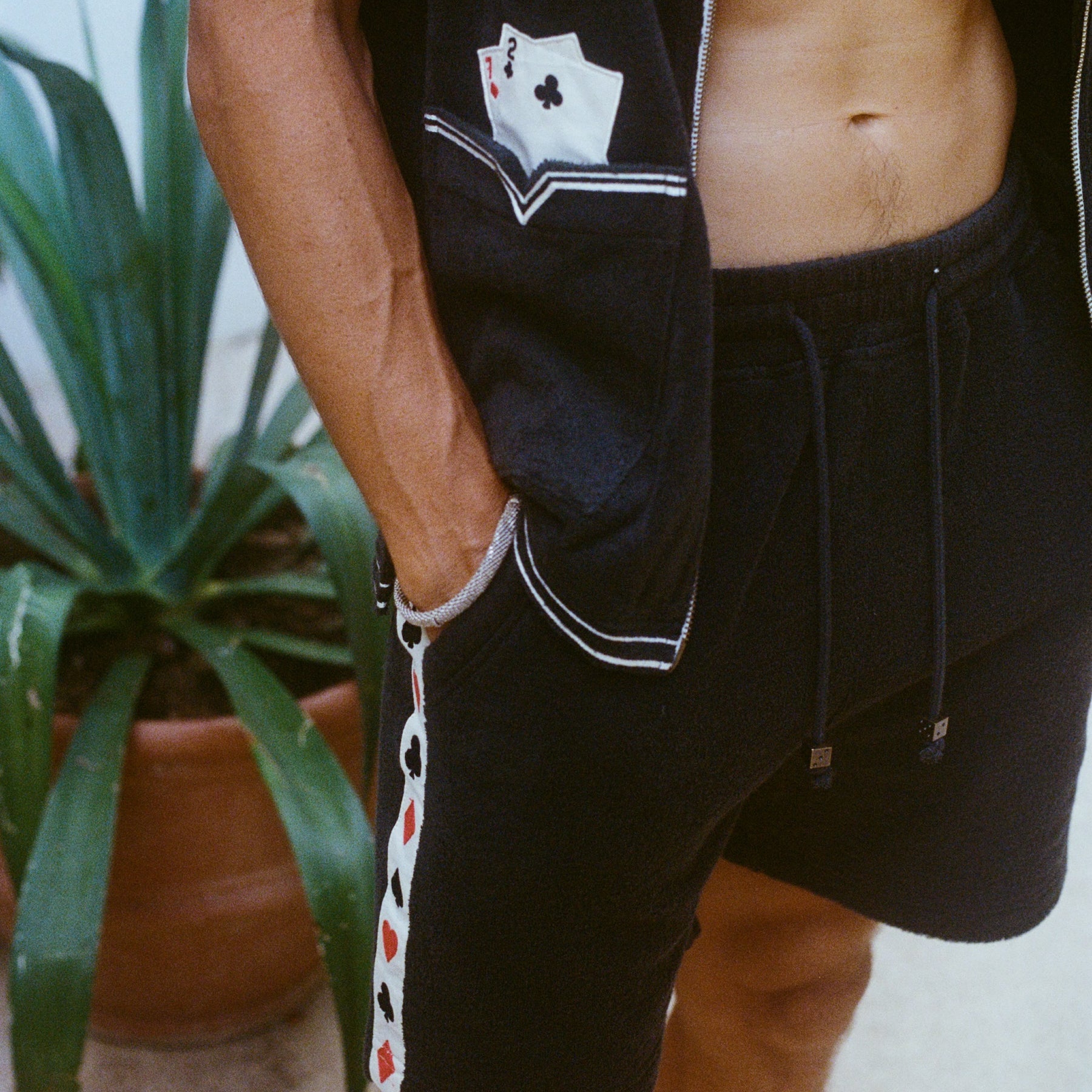 Close-up of a model wearing the green terry cloth shirt and matching shorts. The shirt has an embroidered pocket with two playing cards, and the shorts feature a vertical side stripe patterned with red and black playing card suits. His hand rests in his pocket.