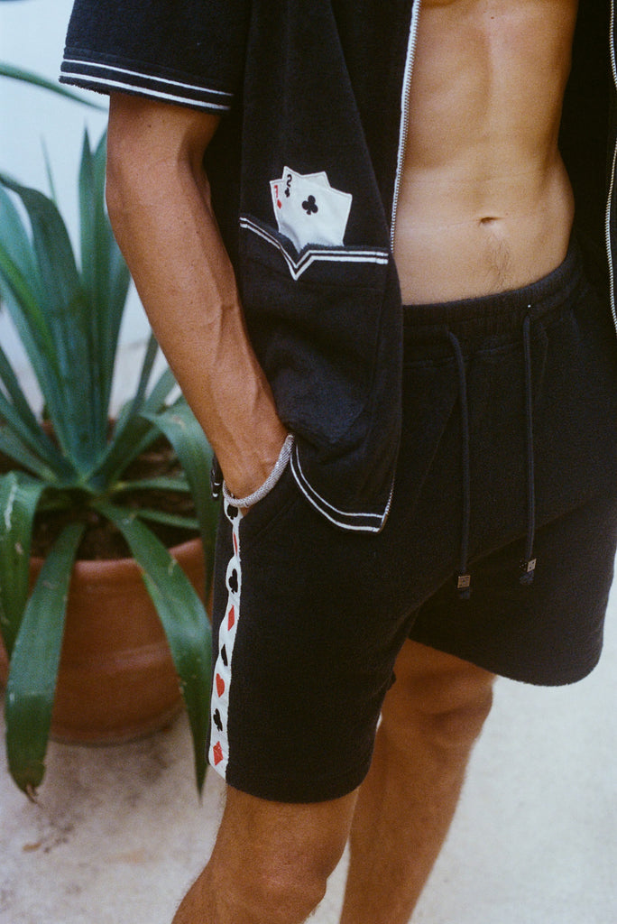 Close-up of a model wearing the green terry cloth shirt and matching shorts. The shirt has an embroidered pocket with two playing cards, and the shorts feature a vertical side stripe patterned with red and black playing card suits. His hand rests in his pocket.