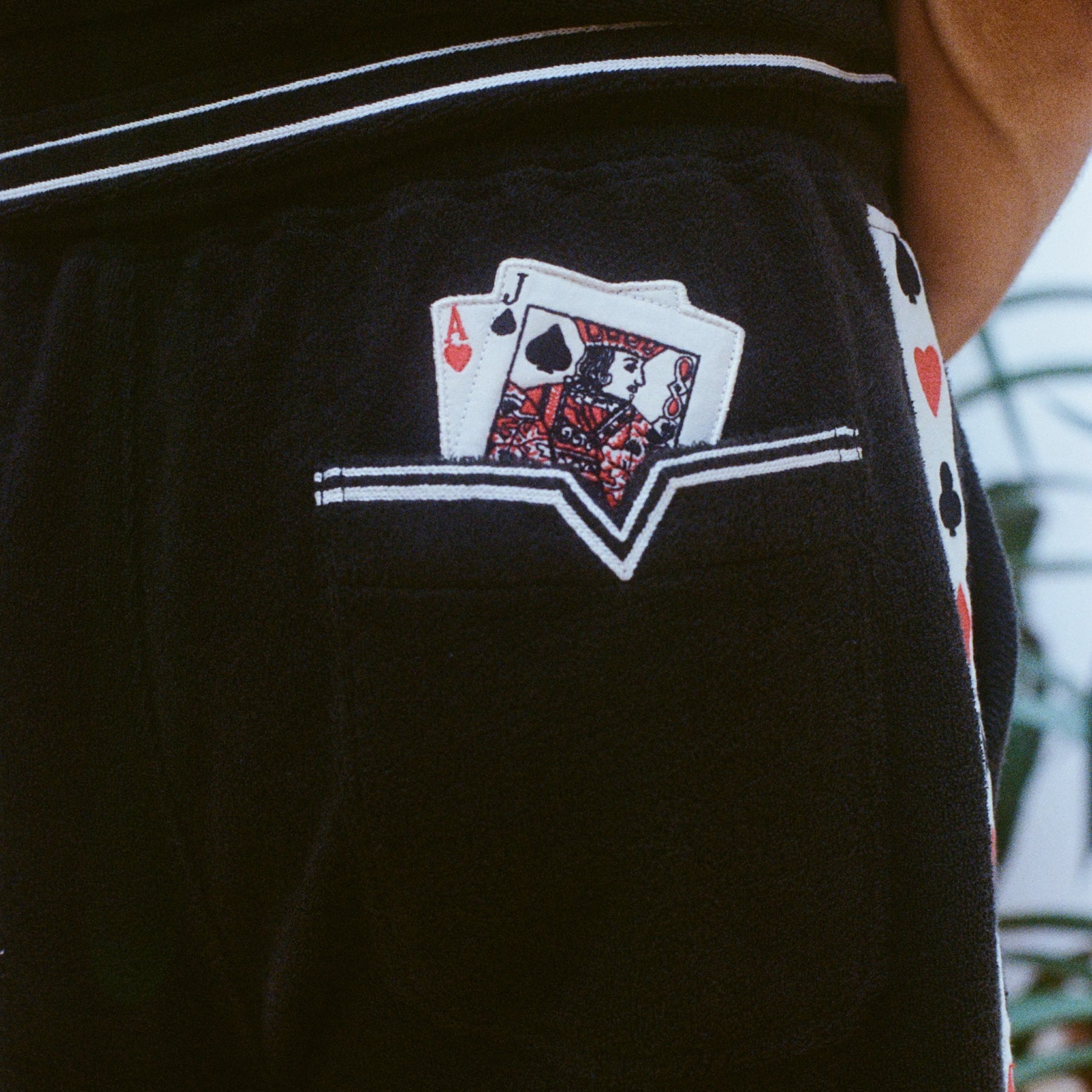 Close-up shot of shorts with playing card patches showing an Ace and Jack of spades in a back pocket, with a white stripe detail and a side panel of playing card suits.