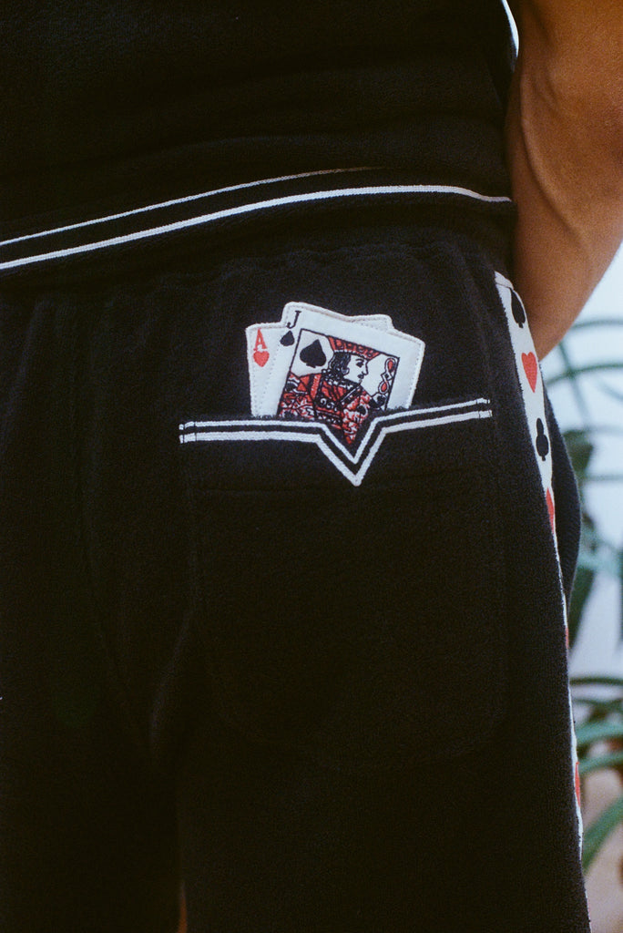 Close-up shot of shorts with playing card patches showing an Ace and Jack of spades in a back pocket, with a white stripe detail and a side panel of playing card suits.