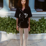 Female model wearing the black embroidered short-sleeve shirt and cream open-knit pants with black sandals. 