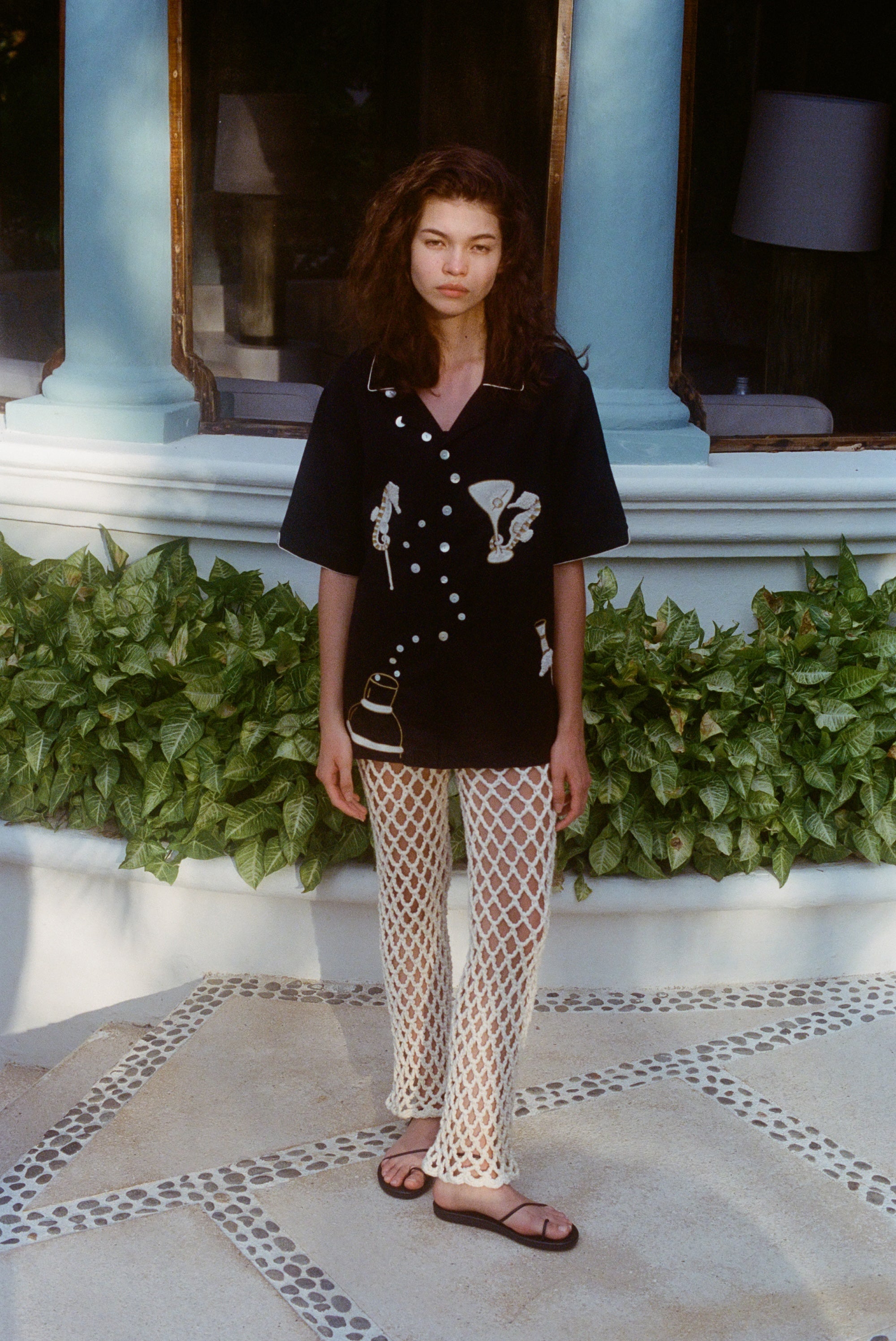 Female model wearing the black embroidered short-sleeve shirt and cream open-knit pants with black sandals. 