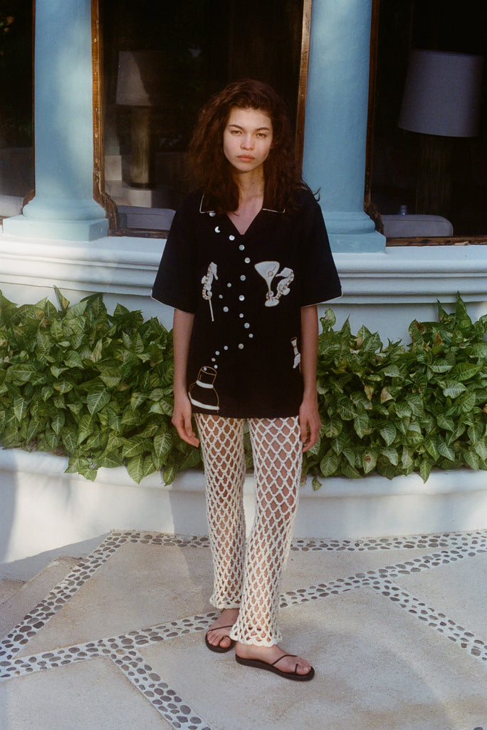 Female model wearing the black embroidered short-sleeve shirt and cream open-knit pants with black sandals. 