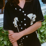 Close-up of a woman standing in front of greenery, wearing the black short-sleeve shirt with embroidered seahorses and barware. Her arms are crossed gently in front of her, and her face is partially out of frame.
