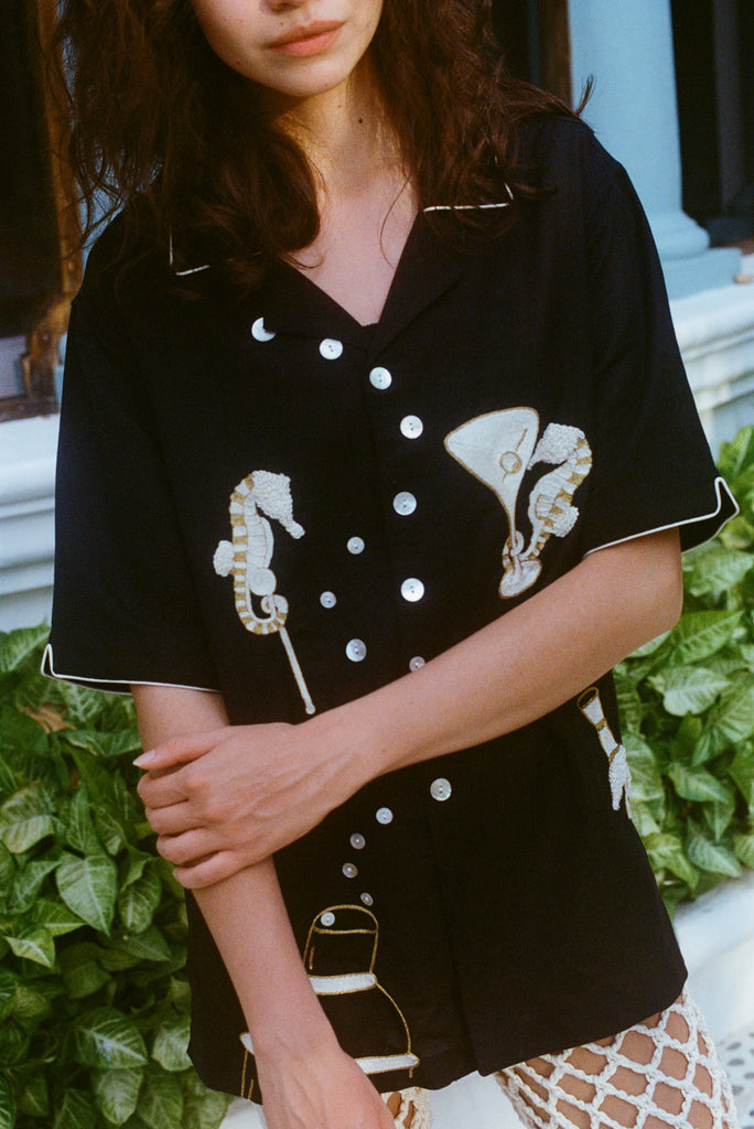 Close-up of a woman standing in front of greenery, wearing the black short-sleeve shirt with embroidered seahorses and barware. Her arms are crossed gently in front of her, and her face is partially out of frame.
