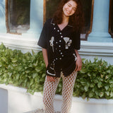 Female model smiling with her eyes closed in Black button down shirt with cream open knit pants and black sandals. 