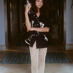 Woman standing in a shaded archway holding a wine glass, wearing a black embroidered short-sleeve shirt and cream open-knit pants with black sandals. Her expression is calm and composed, with arms loosely crossed.