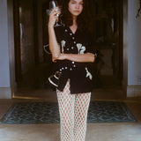 Woman standing in a shaded archway holding a wine glass, wearing a black embroidered short-sleeve shirt and cream open-knit pants with black sandals. Her expression is calm and composed, with arms loosely crossed.