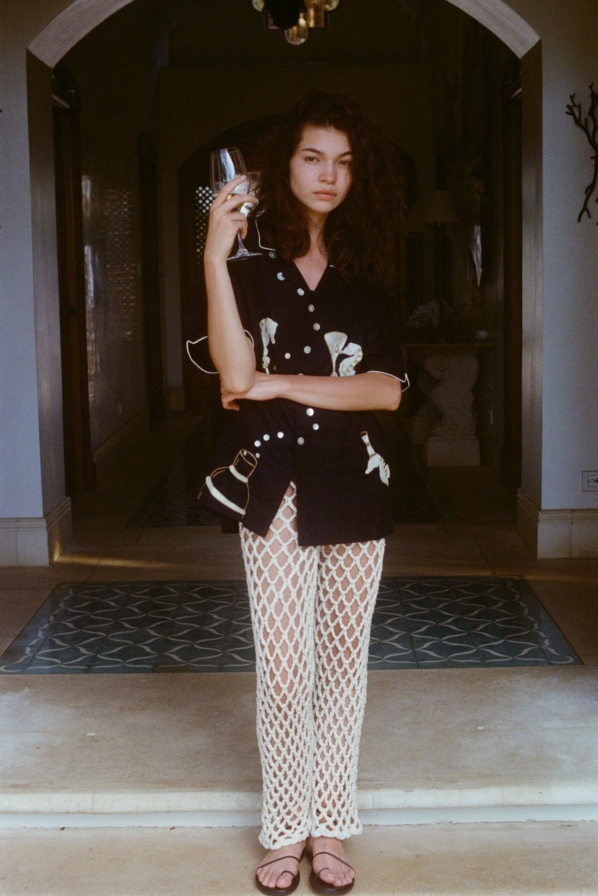 Woman standing in a shaded archway holding a wine glass, wearing a black embroidered short-sleeve shirt and cream open-knit pants with black sandals. Her expression is calm and composed, with arms loosely crossed.