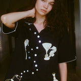 Close up of female model with her arm behind her neck, showing off the black button down shirt and gazing into camera. 