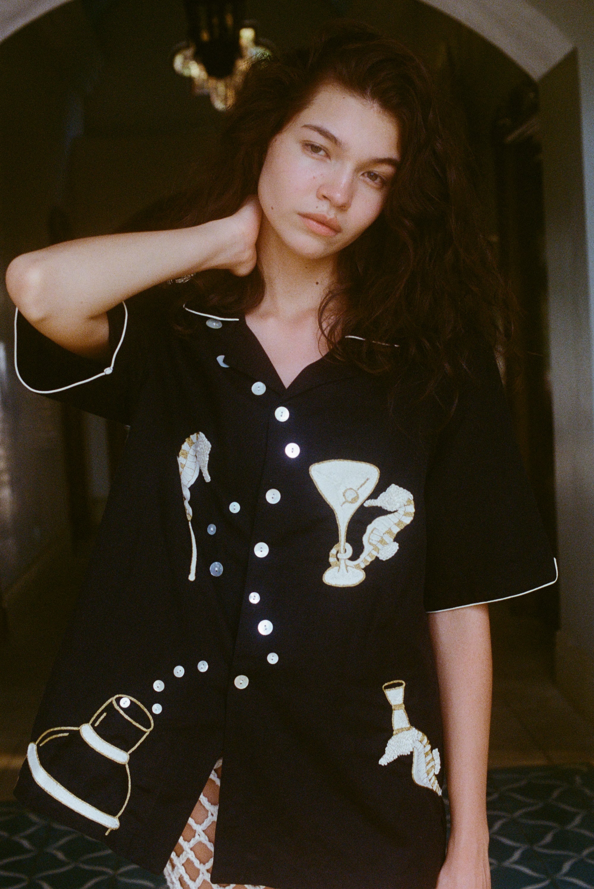 Close up of female model with her arm behind her neck, showing off the black button down shirt and gazing into camera. 