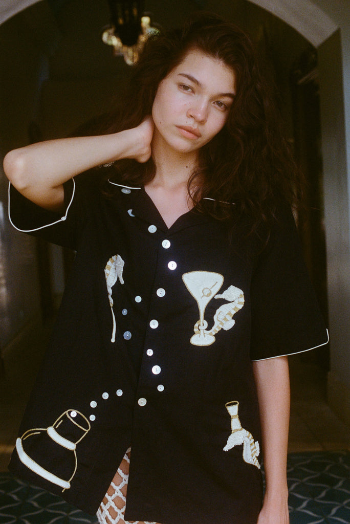 Close up of female model with her arm behind her neck, showing off the black button down shirt and gazing into camera. 