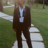 Man wearing a tuxedo jacket, untied bow tie, and relaxed black drawstring pants, standing barefoot.