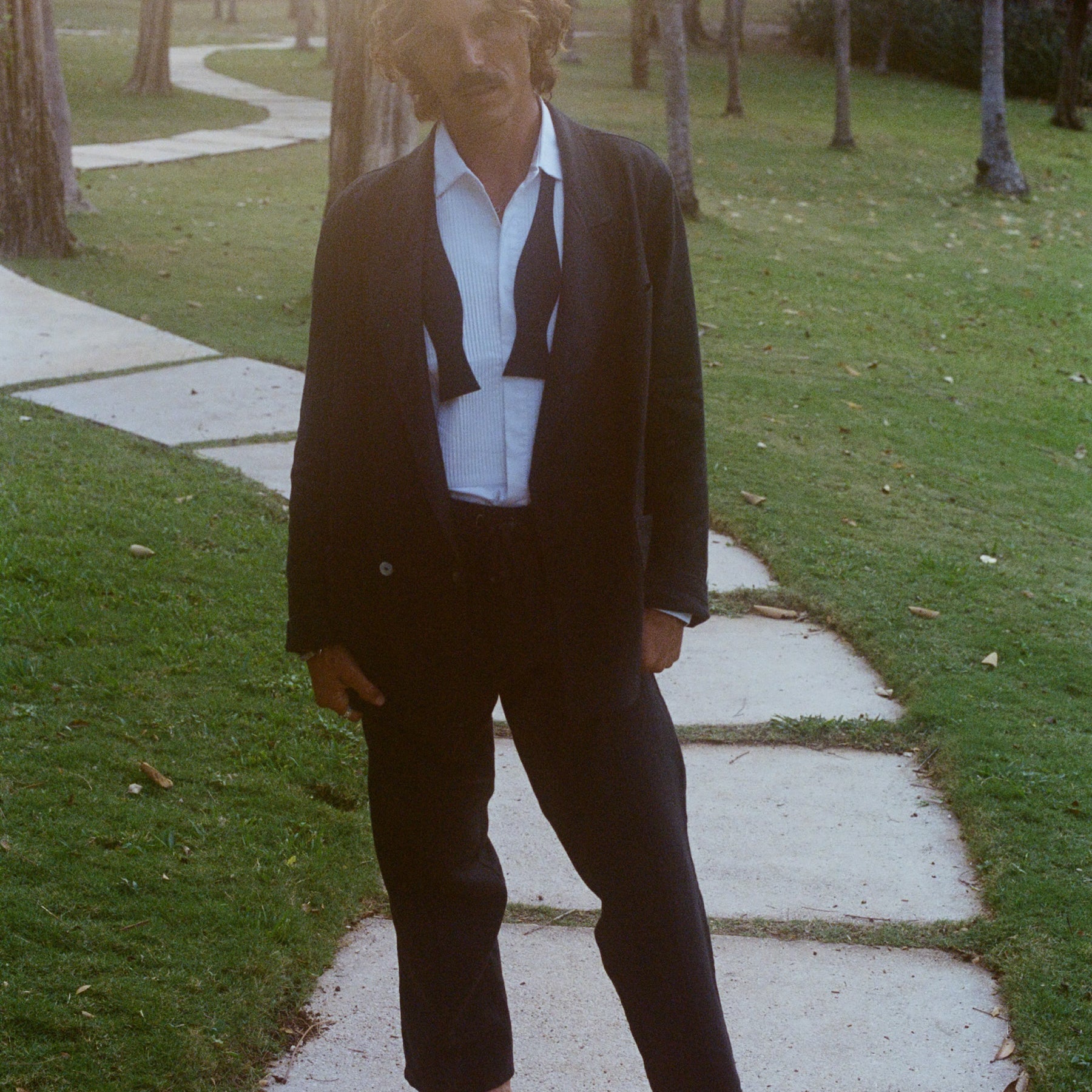 Man wearing a tuxedo jacket, untied bow tie, and relaxed black drawstring pants, standing barefoot.