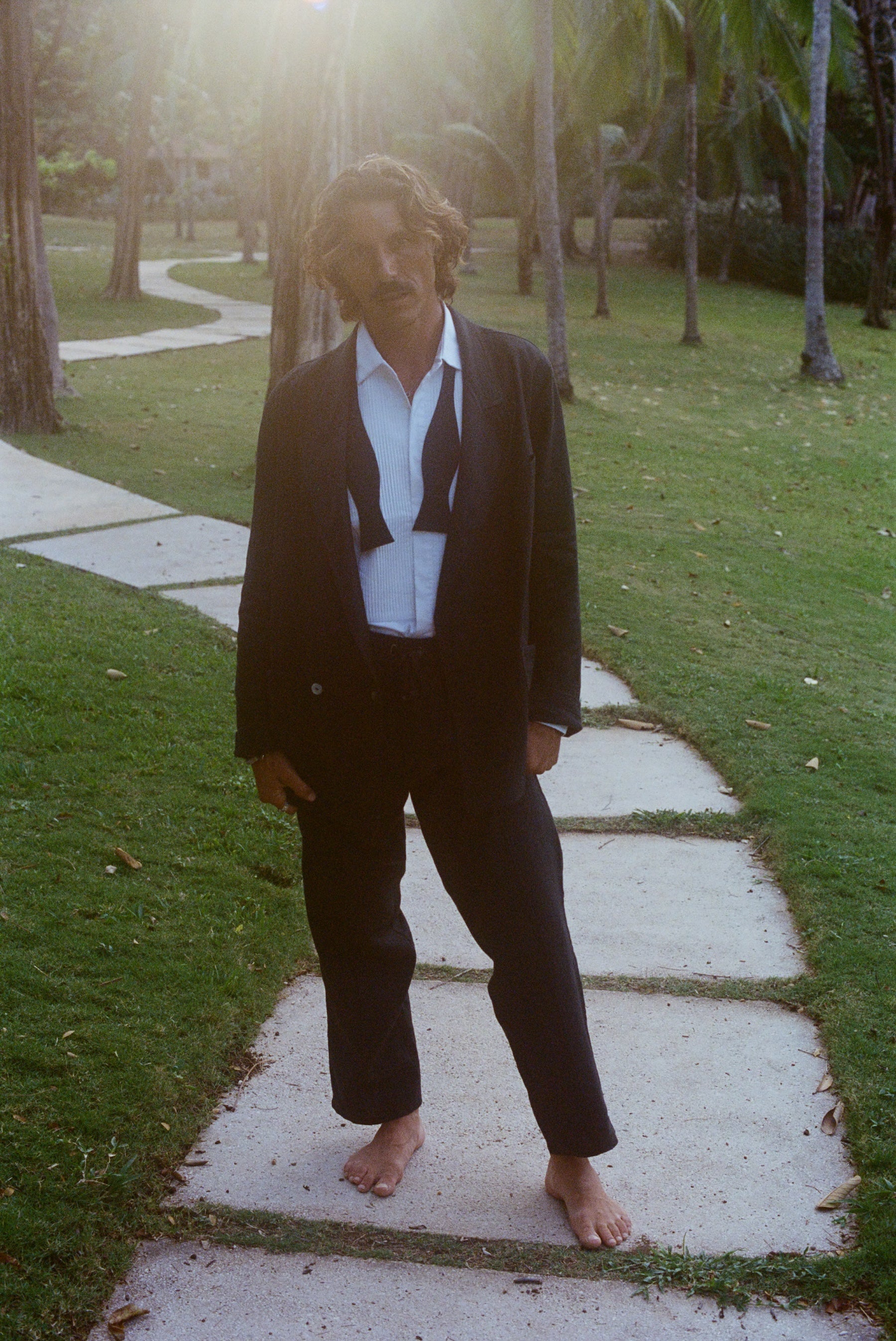 Man wearing a tuxedo jacket, untied bow tie, and relaxed black drawstring pants, standing barefoot.