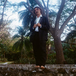 Man wearing the Easy-Going Linen Tuxedo, standing on a stone wall.  His hands are holding the ends of an untied bowtie. 