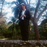 Man wearing the Easy-Going Linen Tuxedo, standing on a stone wall.  His hands are holding the ends of an untied bowtie. 