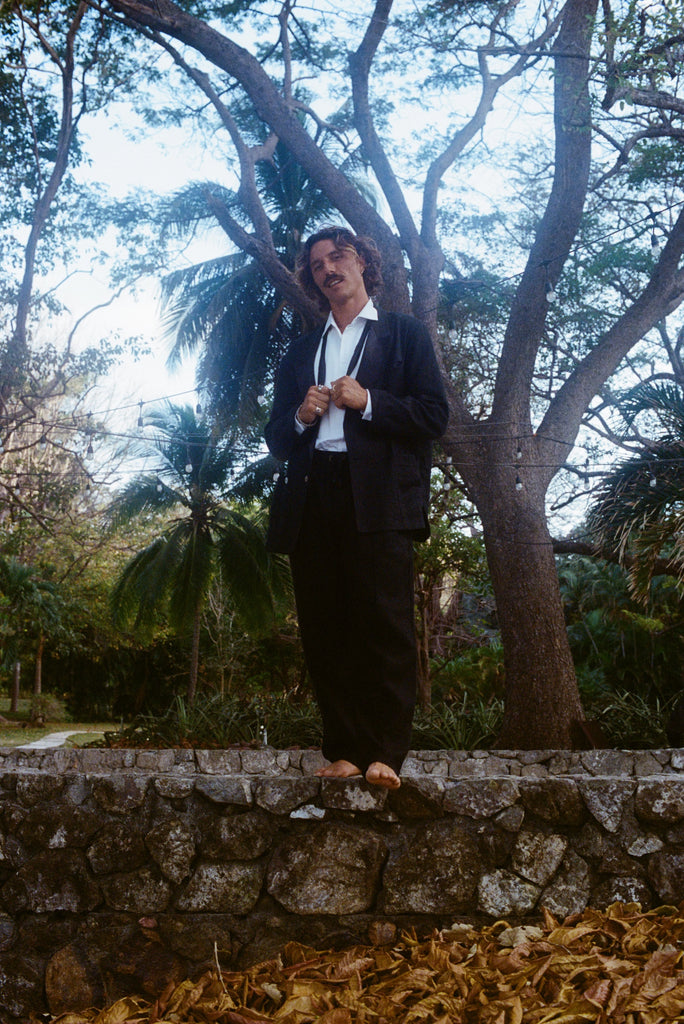 Man wearing the Easy-Going Linen Tuxedo, standing on a stone wall.  His hands are holding the ends of an untied bowtie. 