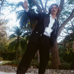 Man dressed in a tuxedo jacket, white shirt, and black drawstring pants, standing barefoot on a stone ledge and raising a glass of white wine.