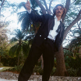 Man dressed in a tuxedo jacket, white shirt, and black drawstring pants, standing barefoot on a stone ledge and raising a glass of white wine.