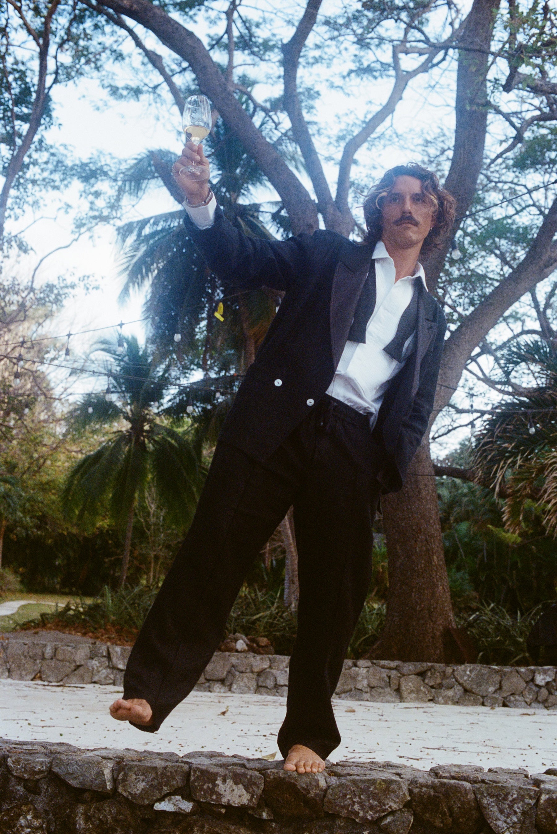 Man dressed in a tuxedo jacket, white shirt, and black drawstring pants, standing barefoot on a stone ledge and raising a glass of white wine.