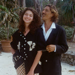 Two people standing outdoors, surrounded by tropical trees, smiling and laughing together. The woman in front wears the black embroidered shirt and cream open-knit pants; the man behind her wears a black suit with an open white-collared shirt. 