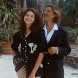 Two people standing outdoors, surrounded by tropical trees, smiling and laughing together. The woman in front wears the black embroidered shirt and cream open-knit pants; the man behind her wears a black suit with an open white-collared shirt. 