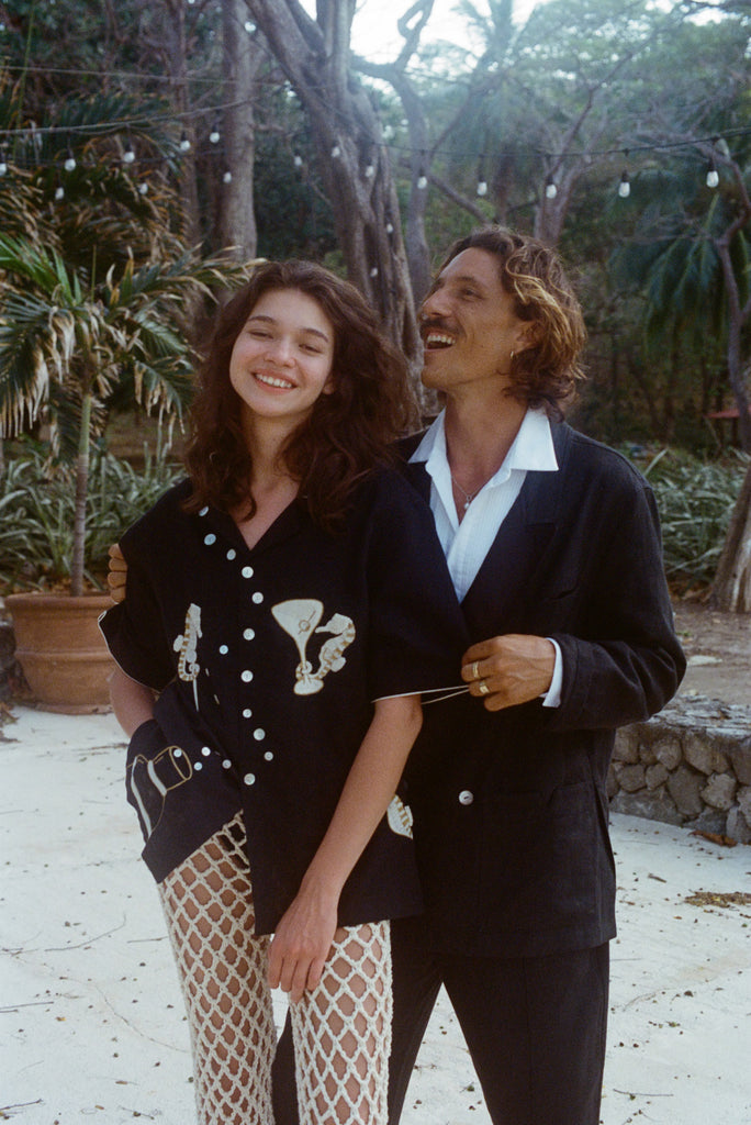 Two people standing outdoors, surrounded by tropical trees, smiling and laughing together. The woman in front wears the black embroidered shirt and cream open-knit pants; the man behind her wears a black suit with an open white-collared shirt. 
