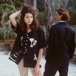 Woman standing outdoors with one hand in her hair, wearing a black embroidered shirt and cream open-knit pants. A man in a black suit stands behind her, facing away. The setting is tropical, with palm trees, string lights, and sandy ground.